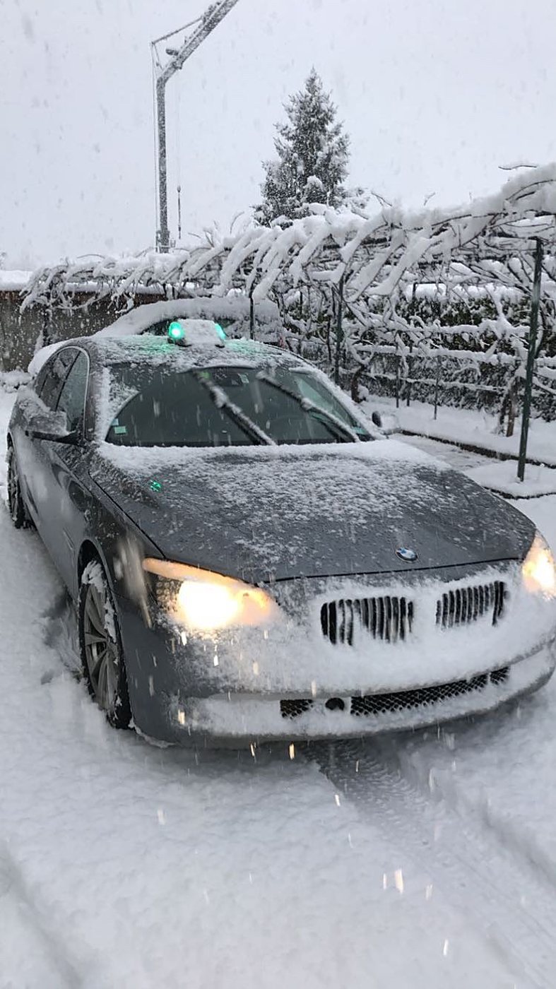 Une berline grise recouverte de neige, phares allumés et lumignon vert sur le toit, stationnée dans une allée enneigée pendant une chute de neige.
