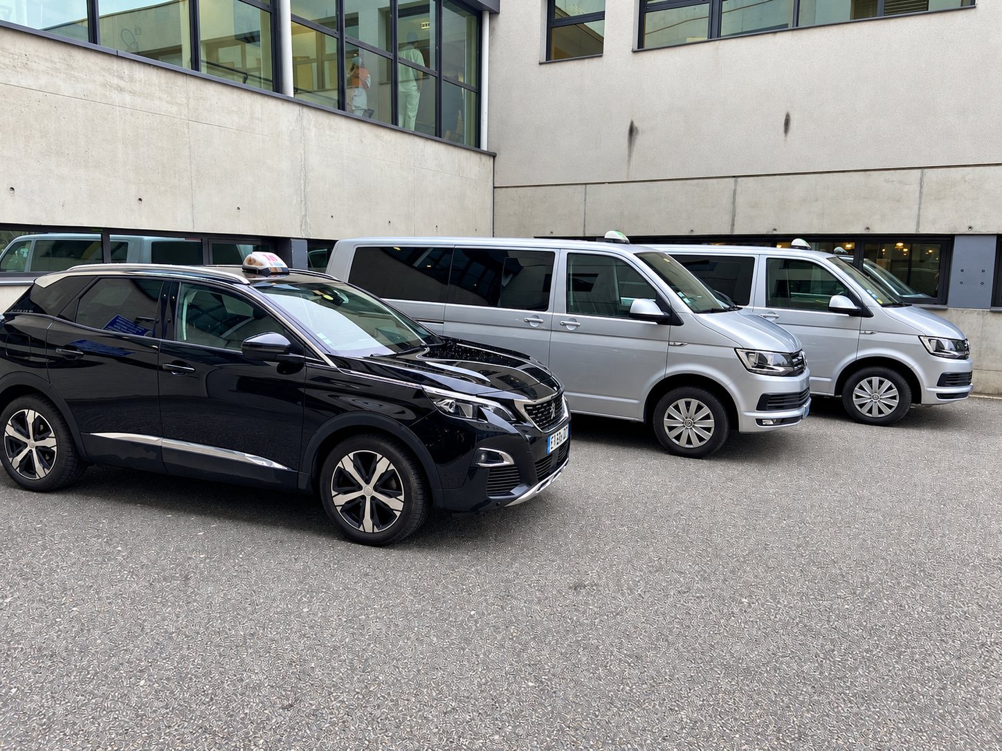 Un SUV noir et deux minibus gris équipés de enseignes de taxi sont garés devant un bâtiment en béton.