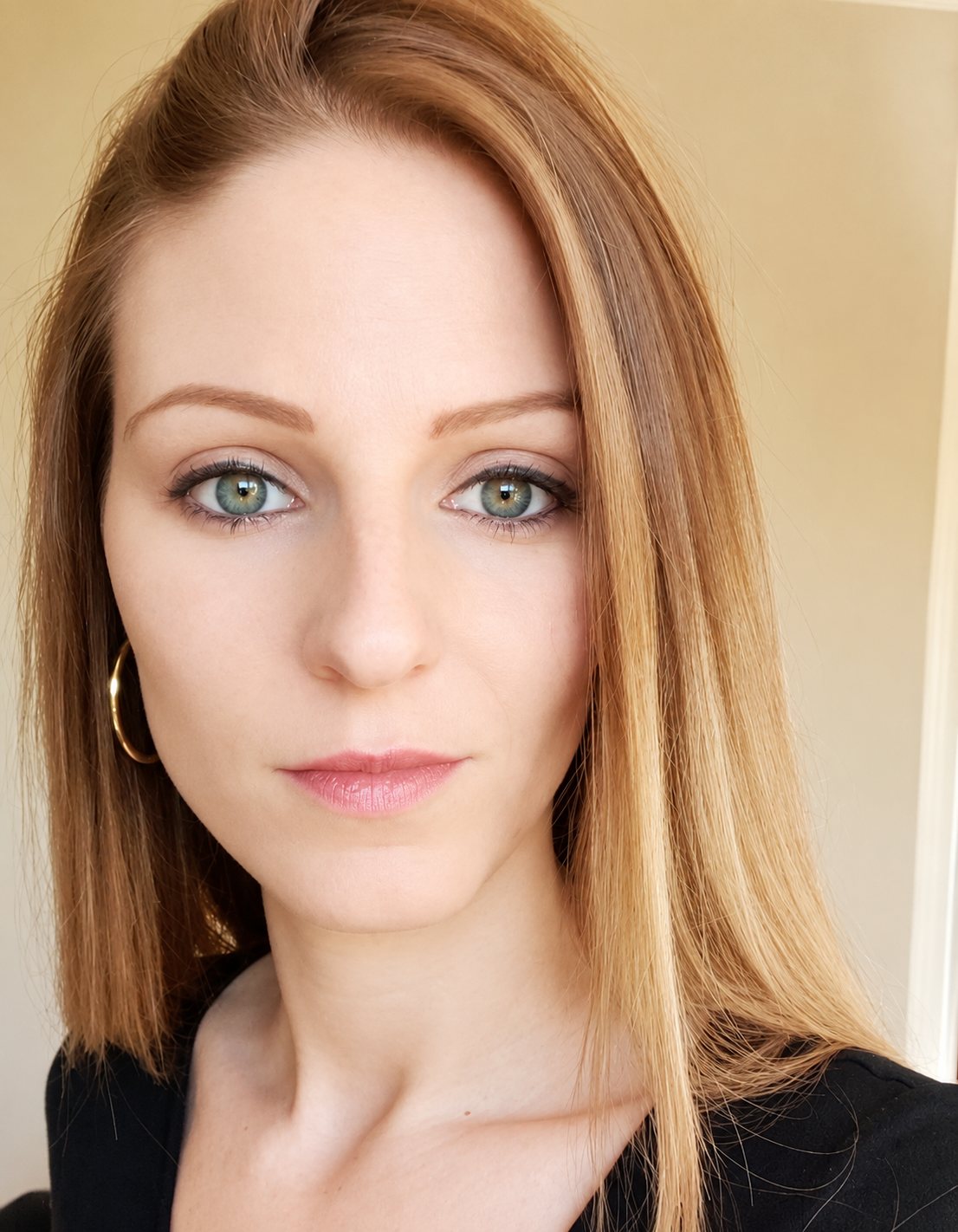 Portrait en gros plan d'une jeune femme aux yeux clairs et aux longs cheveux lisses roux, portant une boucle d'oreille dorée et un haut noir, devant un mur beige.