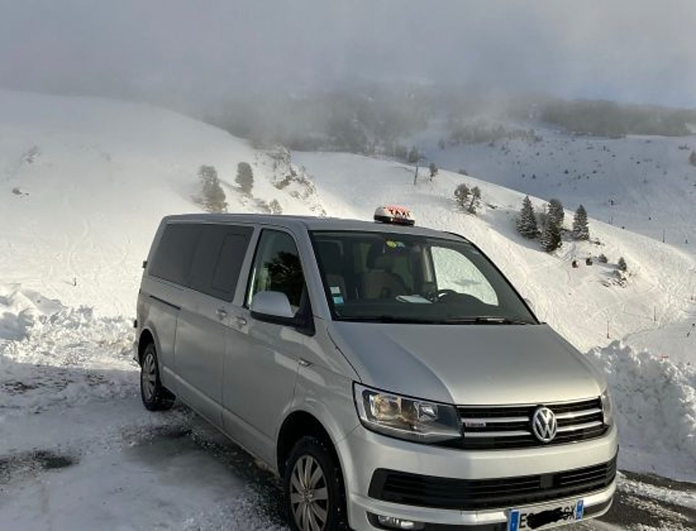 Un minibus Volkswagen gris équipé d'un lumineux de taxi sur le toit est garé sur une route de montagne enneigée, sous un ciel brumeux.