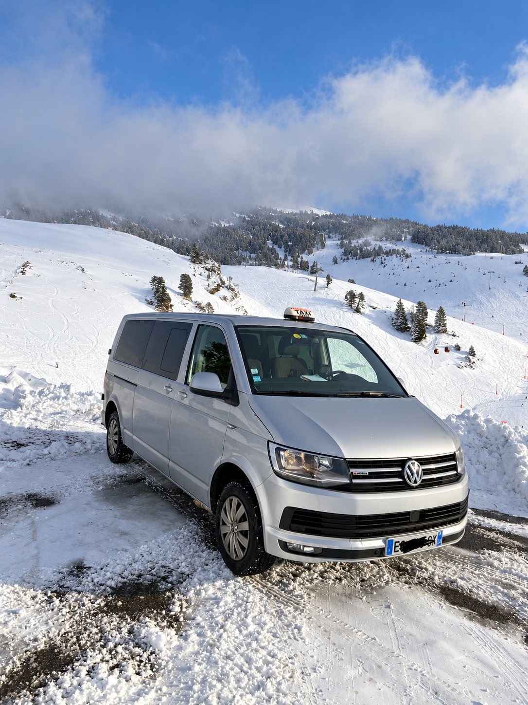 Un minibus Volkswagen gris équipé d'un lumineux de taxi est garé sur une route enneigée, face à une pente de ski dans la brume.