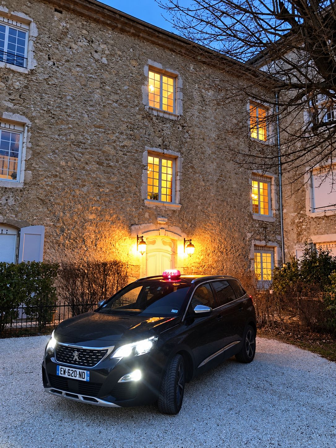 Un taxi Peugeot sombre avec son lumineux allumé est garé sur le gravier, au crépuscule, devant une grande bâtisse en pierre aux fenêtres éclairées.