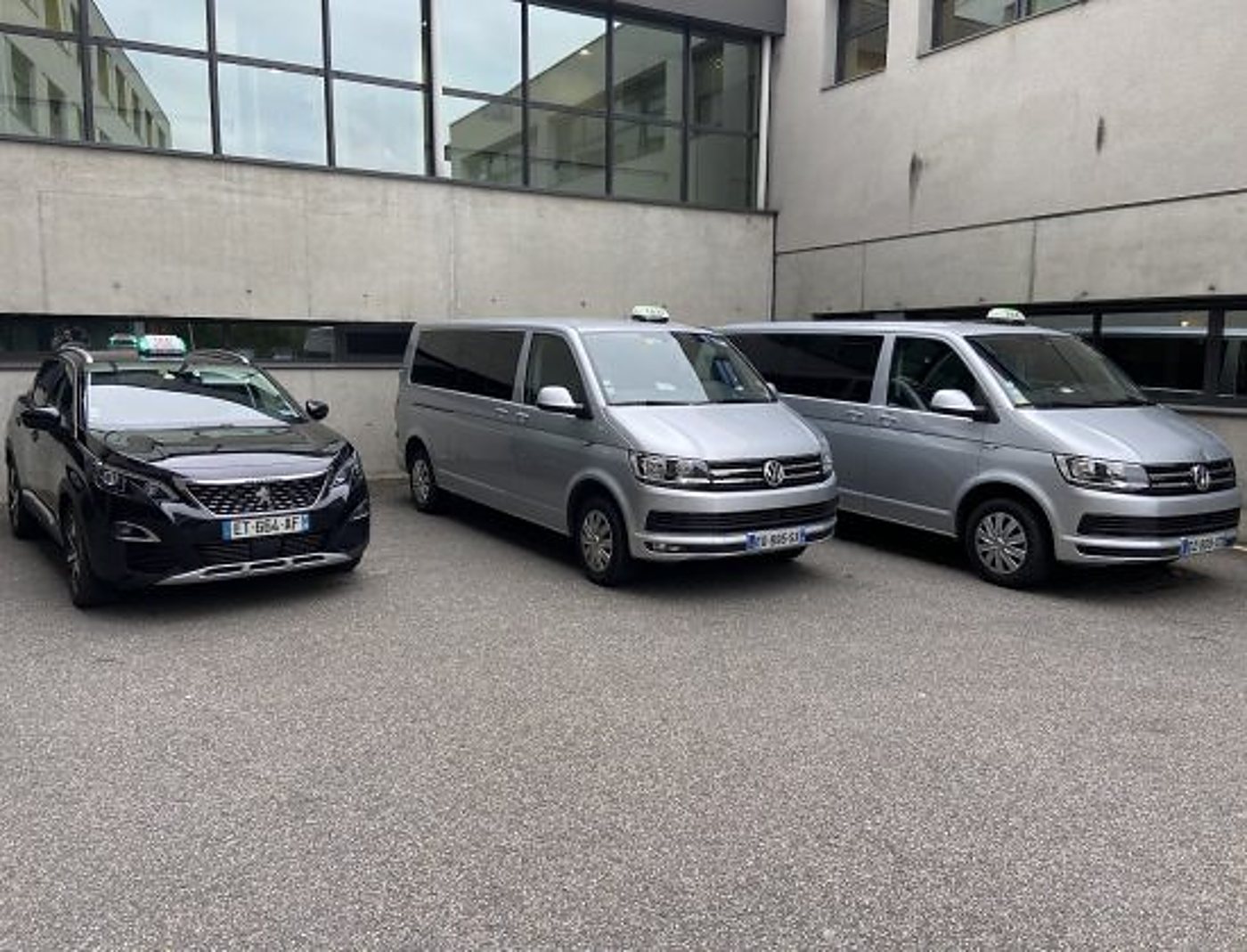 Un SUV Peugeot noir et deux minibus Volkswagen gris avec enseignes de taxi sont garés côte à côte devant un bâtiment en béton.