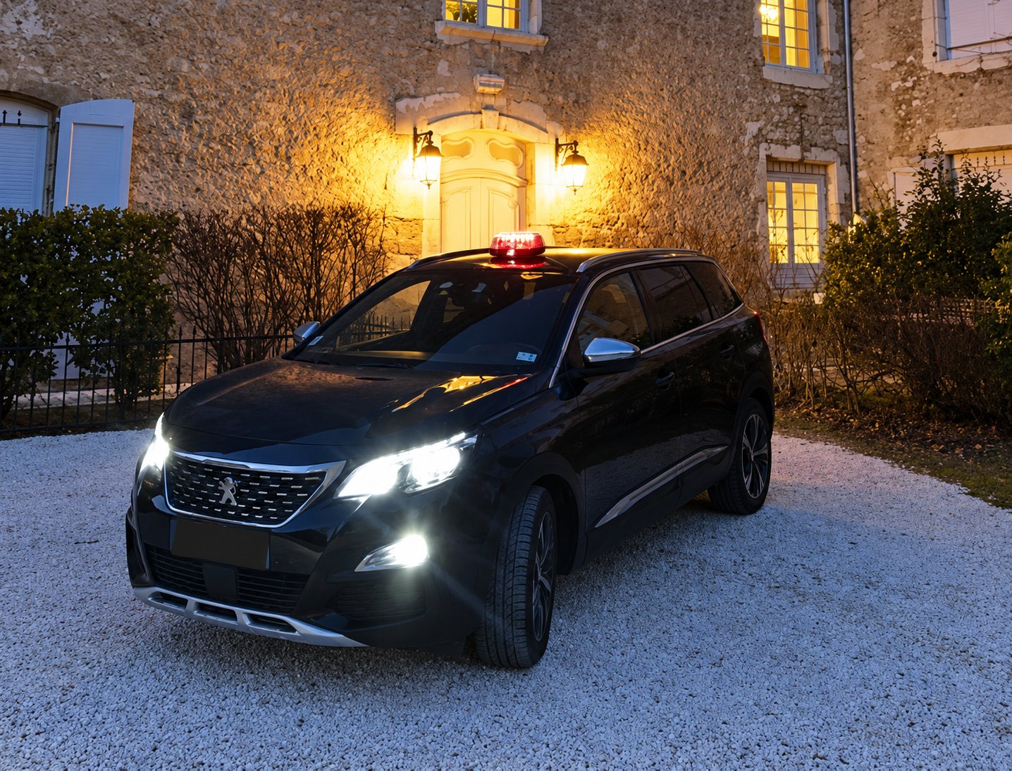 Un SUV Peugeot sombre avec un lumineux rouge sur le toit, phares allumés, garé sur le gravier devant une maison en pierre éclairée au crépuscule.